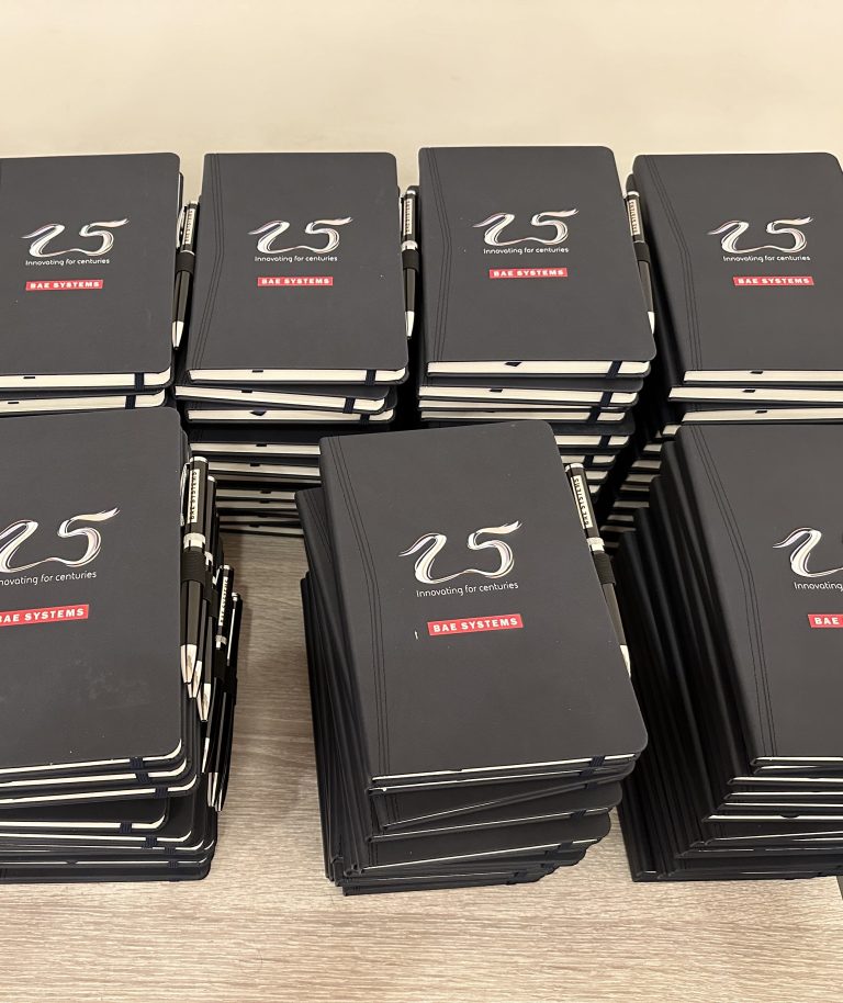 Piles of black BAE System 25th Anniversary branded notebooks